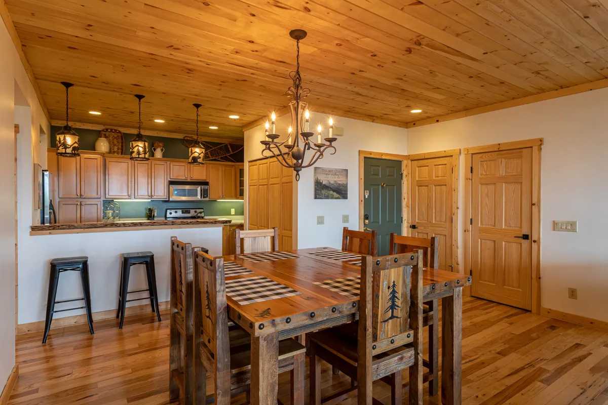 A welcoming dining space with a rustic wooden table and matching bar stools, illuminated by a charming chandelier. The area opens toward the kitchen, with cabinetry visible in the background, suggesting communal meals and gatherings.