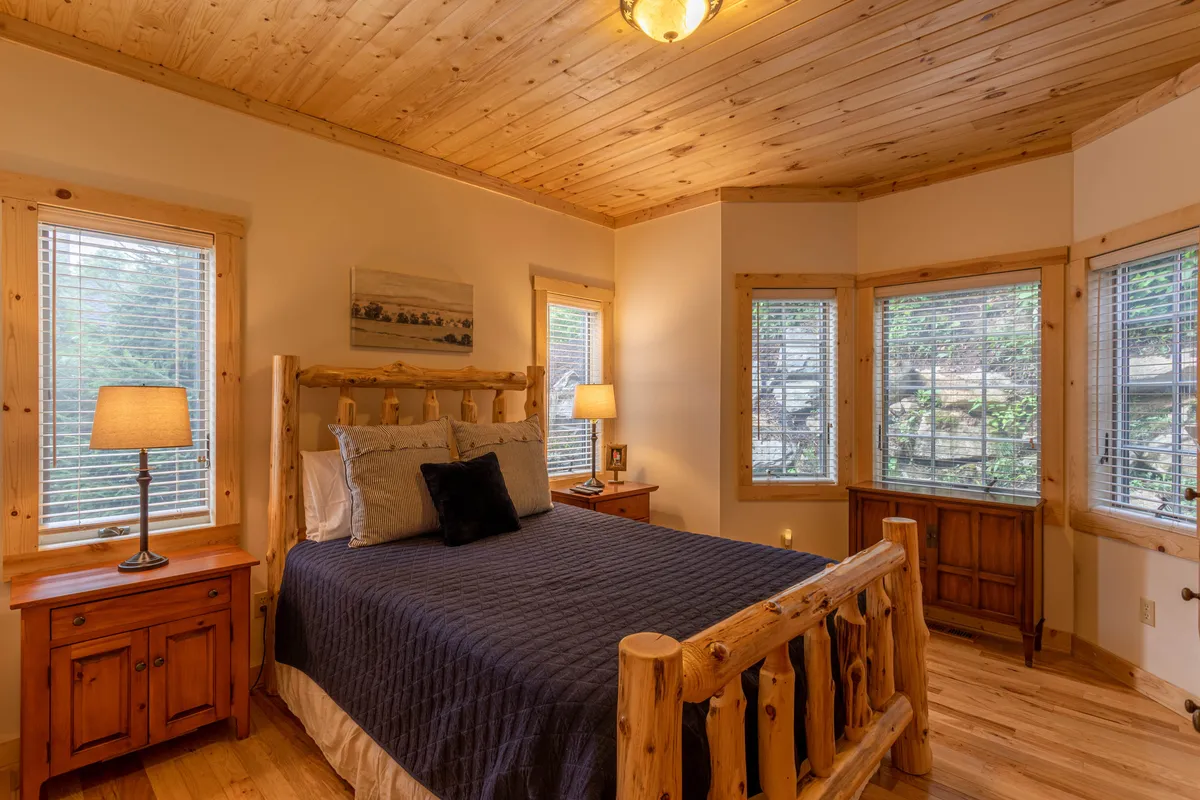 A tranquil master suite featuring a rustic log bed, nightstand with lamp, wooden furniture, and large windows that frame the natural light. The space exudes comfort, making it ideal for restful nights and leisurely mornings, with a strong sense of privacy and retreat.