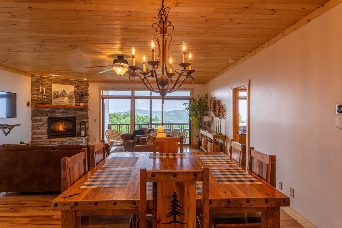 A cozy dining area is featured, framed by large windows that offer expansive mountain views. The fireplace and chandelier add warmth, while the view through the windows visually connects the comfortable interior to the outdoors.