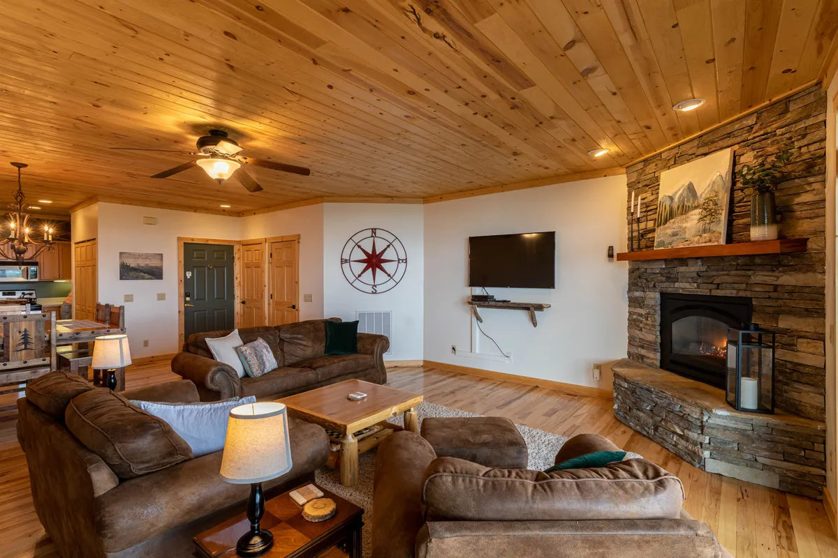A spacious living area featuring a cozy stone fireplace, plush sofa set, a large 55" television, exposed wooden beams on the ceiling, and tasteful decorative wall art. The scene is arranged for warmth and comfort, inviting families to gather for relaxation or movie nights.