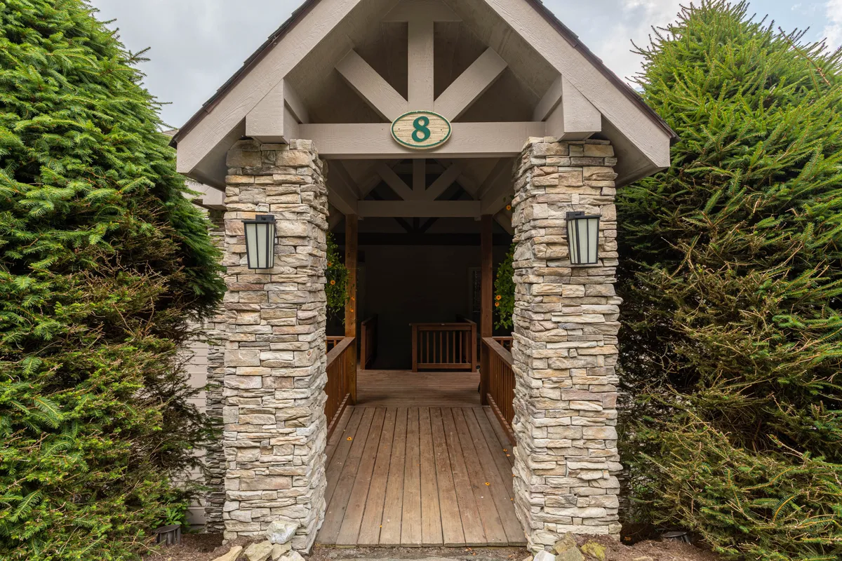 A warm, inviting exterior shot captures stone columns, a wooden walkway, and a decorative ceiling structure, focusing on the building's entrance and house number. The environment feels peaceful and tucked away, reflecting the quiet, private position on the mountain.