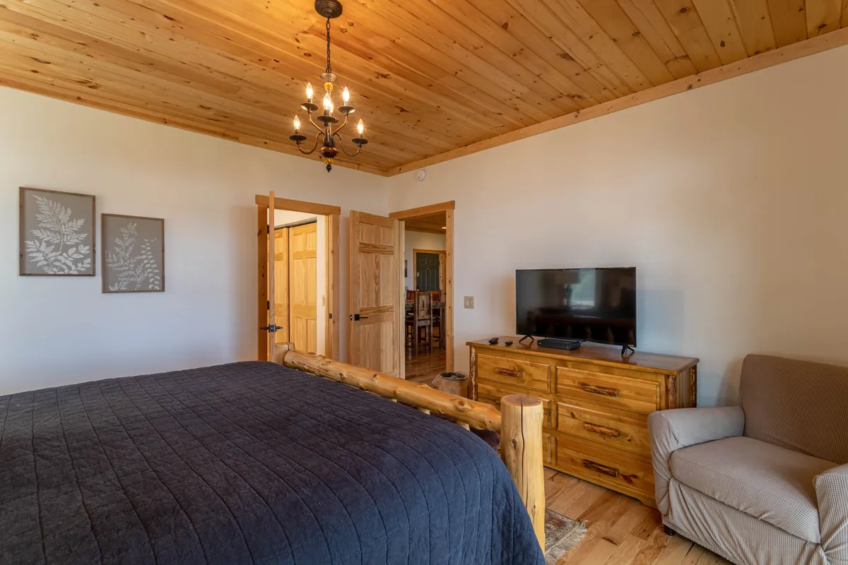 A cozy second bedroom with a classic wooden bed frame, elegant chandelier, television, dresser, and an inviting armchair. The room feels warm and comfortable, ideal for unwinding, with plenty of natural light streaming in.