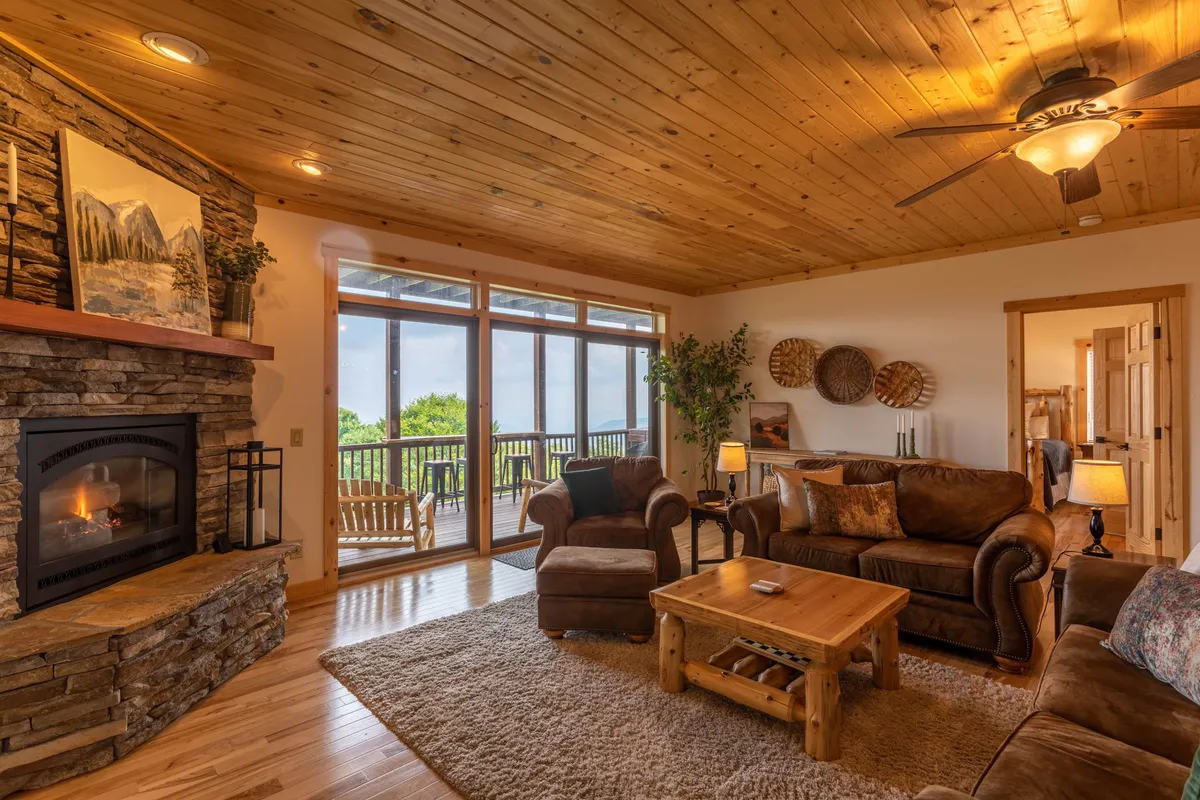 This image showcases the heart of the home's living space: a cozy arrangement of sofas grouped around a rustic gas fireplace, with large windows framing breathtaking mountain views. The inviting atmosphere is perfect for relaxing with family and friends, while the view and glowing fire create a sense of warmth and connection to the outdoors. The sofa set, wooden coffee table, and the 55" TV are visible, emphasizing comfort and entertainment.