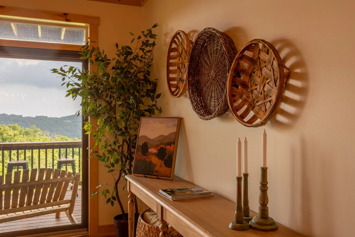 A cozy living area with woven baskets adorning the wall, a green plant adding a touch of nature, framed artwork, a wooden console table, and elegant candlesticks. The space feels warm and inviting, suggesting a perfect spot to unwind or share an intimate moment. Soft morning light filters in, hinting at a view beyond the deck.