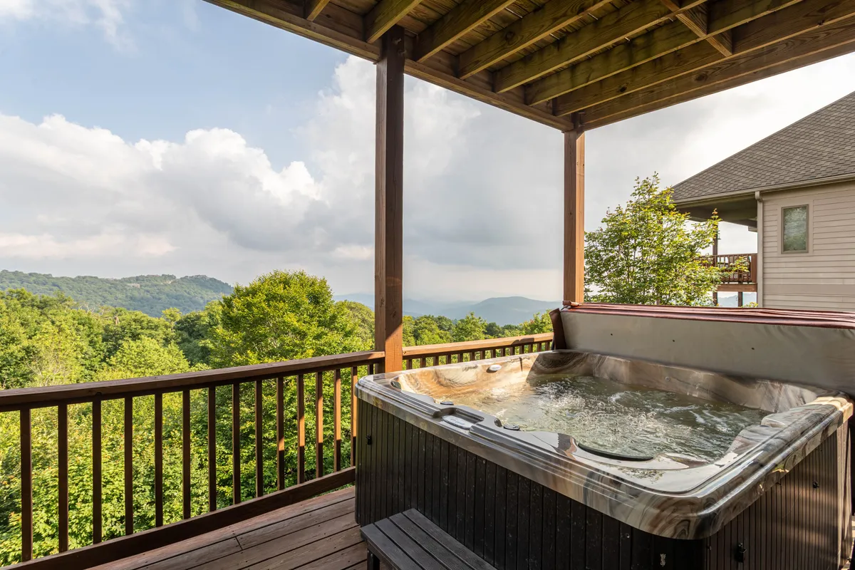 This exterior shot captures the mountain-view deck with a bubbling hot tub prominently placed, framed by sweeping vistas of the Blue Ridge Mountains. The serene setting showcases a peaceful atmosphere perfect for relaxing, with hints of sunrise or starlit skies visible, and a spacious layout ideal for unwinding or socializing.