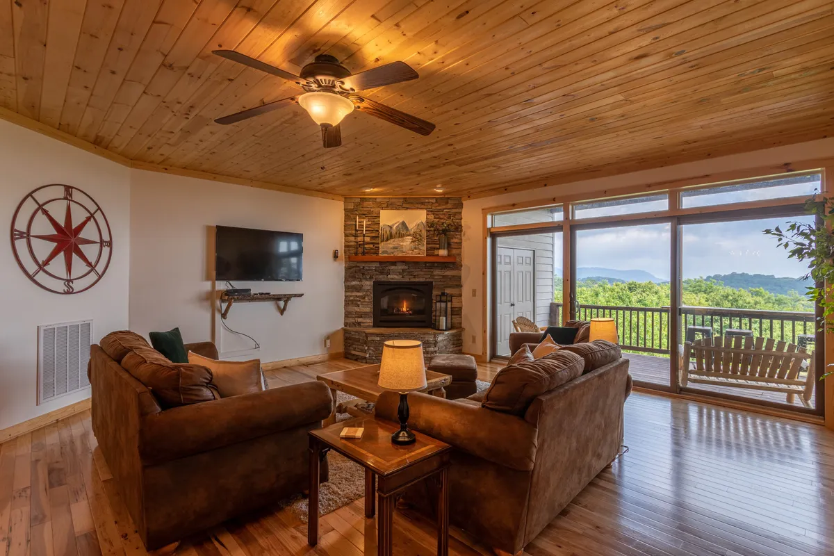 The image showcases a warm, inviting living room with a plush sofa set facing a rustic gas fireplace. Large windows frame breathtaking, long-range mountain views, and a wooden ceiling adds to the cozy, alpine ambiance. The space is perfect for relaxing with family and friends, watching movies, or simply soaking in the sunset after a day on the slopes.
