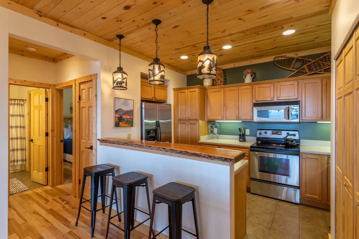 A close-up view of the kitchen area, focusing on the wooden bar counter with pendant lighting, stainless steel appliances, and warm wooden cabinetry. Bar stools invite casual conversation and shared culinary experiences, while the decor adds a touch of homeliness.
