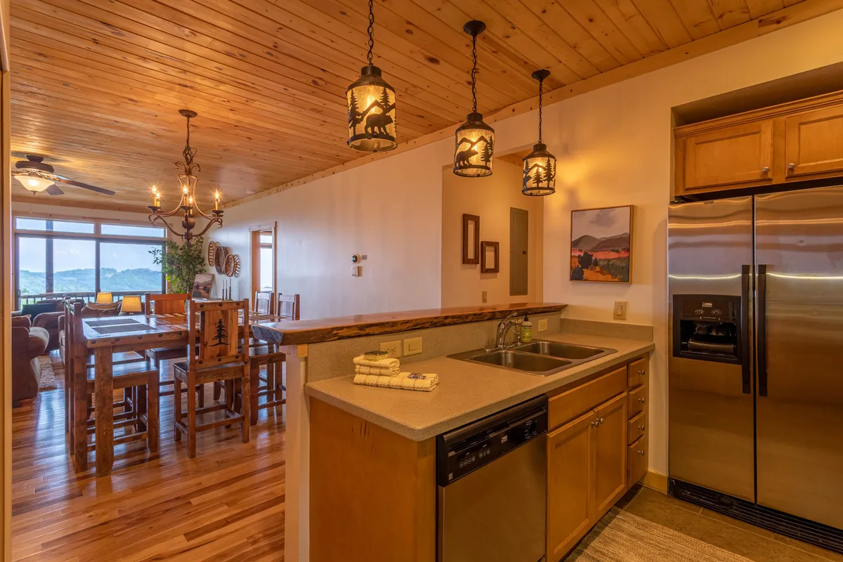 A bright, open perspective featuring the kitchen counter, adjacent dining table, and expansive windows that reveal a panoramic mountain view. Stainless steel appliances, a bar-height counter with stools, and the wooden dining table create a seamless flow between cooking, dining, and socializing—all with a stunning natural backdrop.