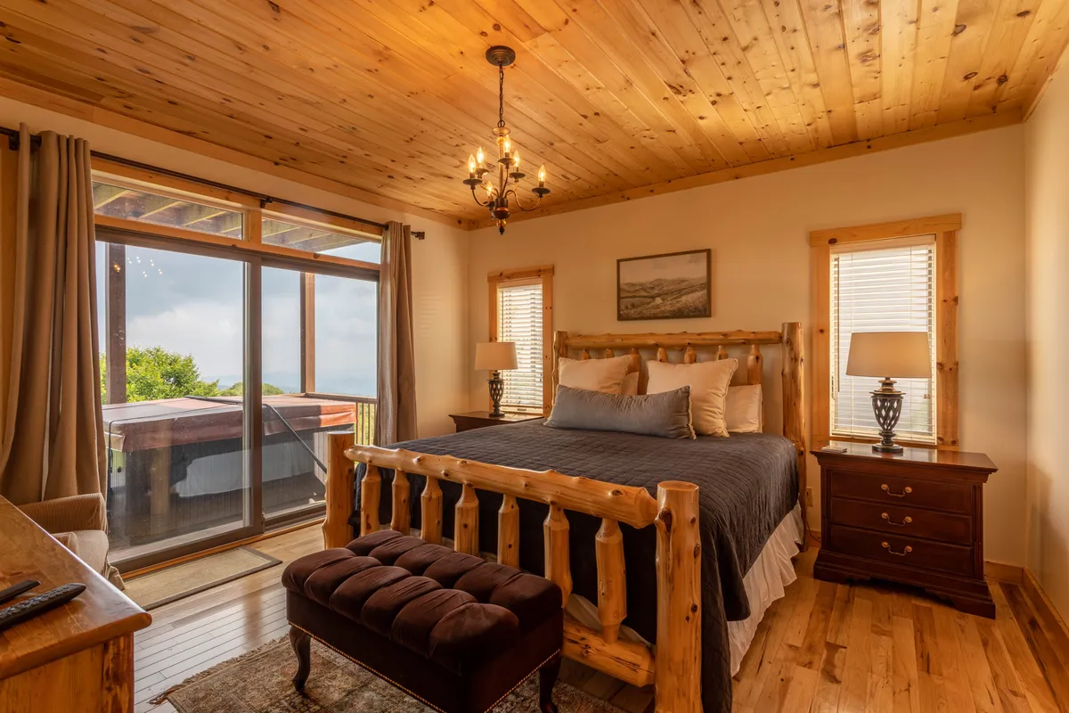 A serene master bedroom featuring a wooden bed frame topped with plush linens, a stylish chandelier, and large windows that frame sweeping mountain views. Nightstands with soft-glow lamps flank the bed, while the calming decor in neutral tones exudes comfort and relaxation.